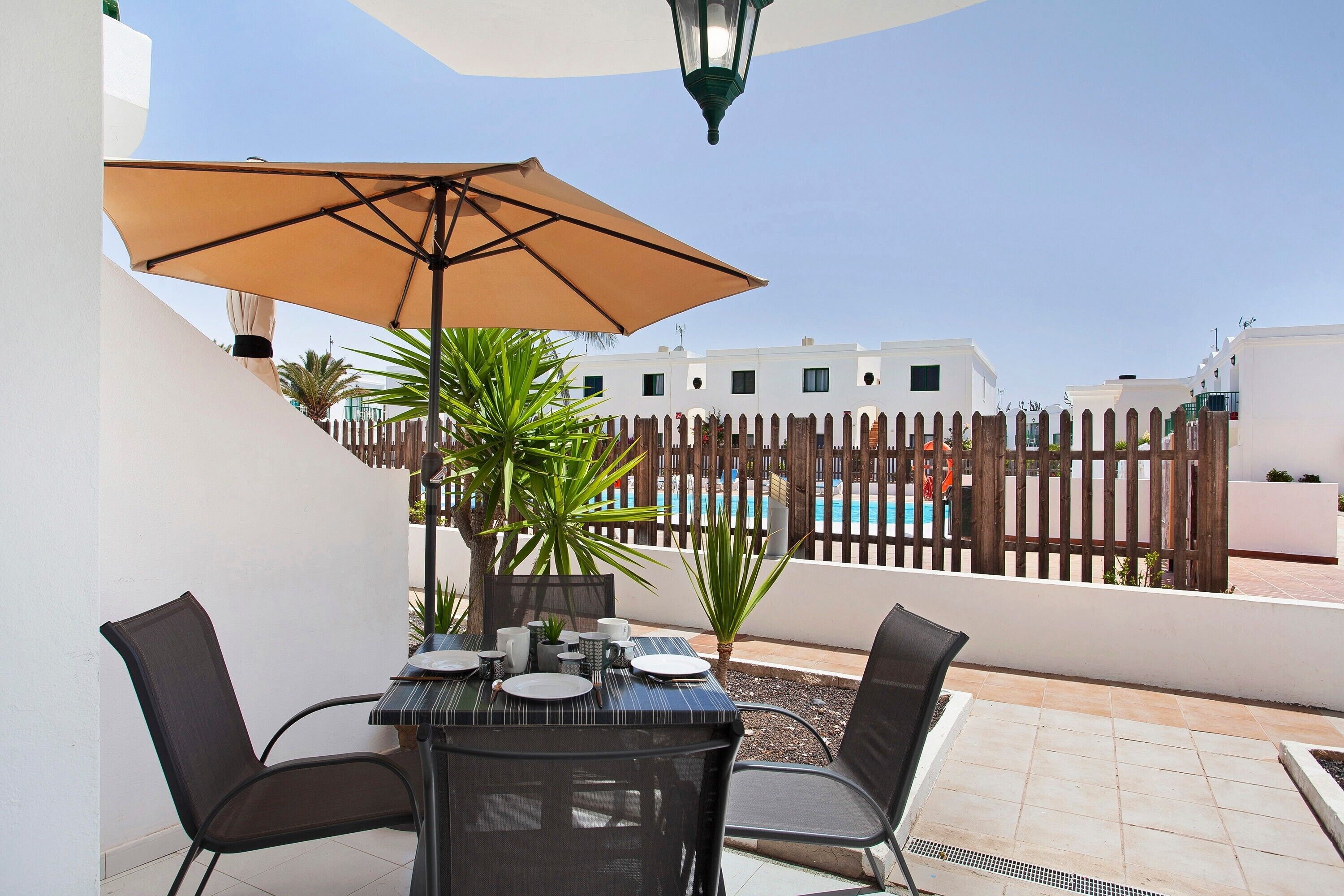 Une terrasse avec une table dressée pour deux personnes, un parasol et une vue sur une piscine et des bâtiments en arrière-plan.