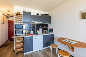 A compact kitchen with a microwave, toaster, and small refrigerator. There is a wooden table with two stools and a wall-mounted shelf above the counter.