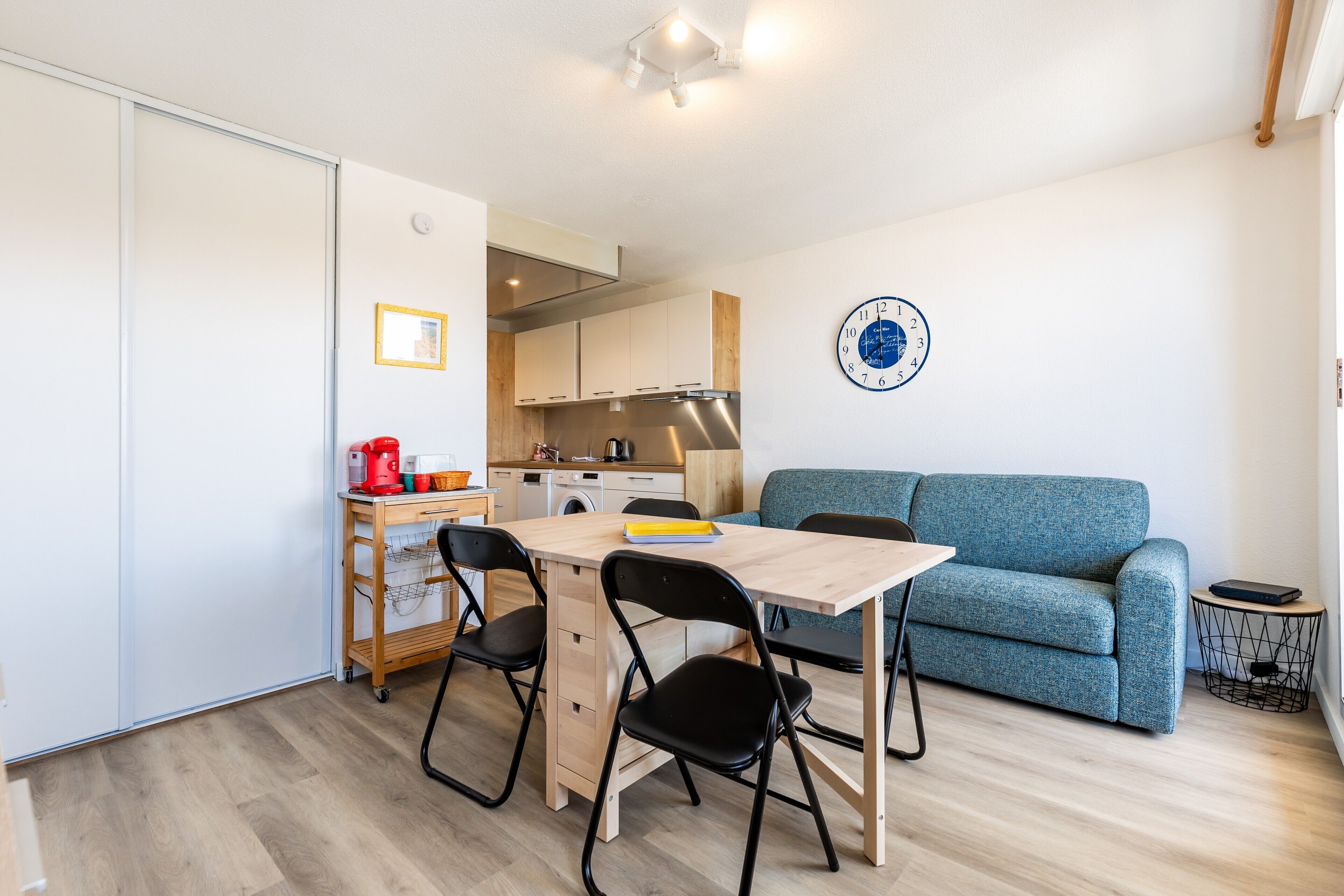 Un espacio habitable compacto con kitchenette, una mesa de comedor con sillas, un sofá y un reloj de pared.