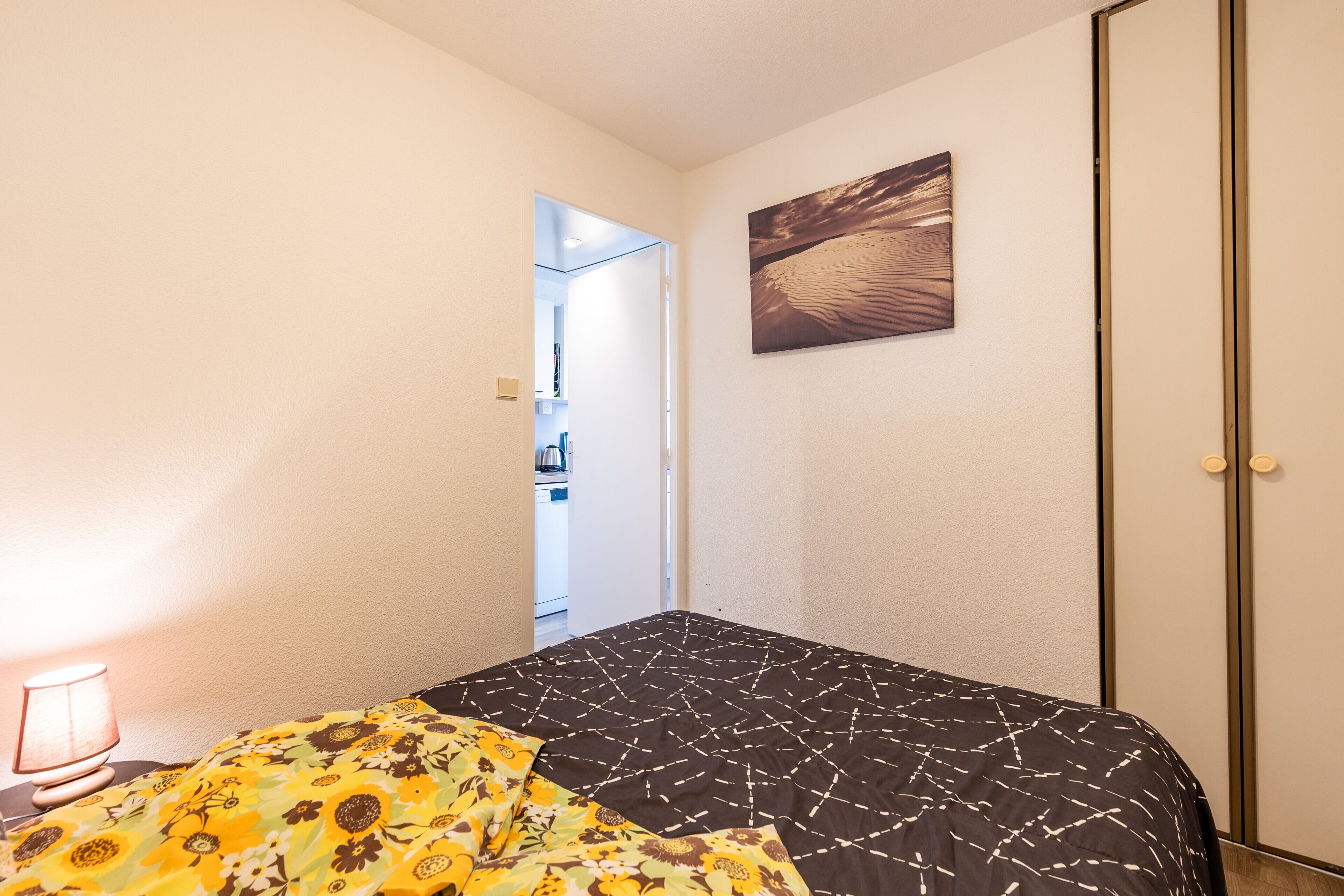 Een slaapkamer met een bed, nachtkastje, lamp, schilderij en een kledingkast.