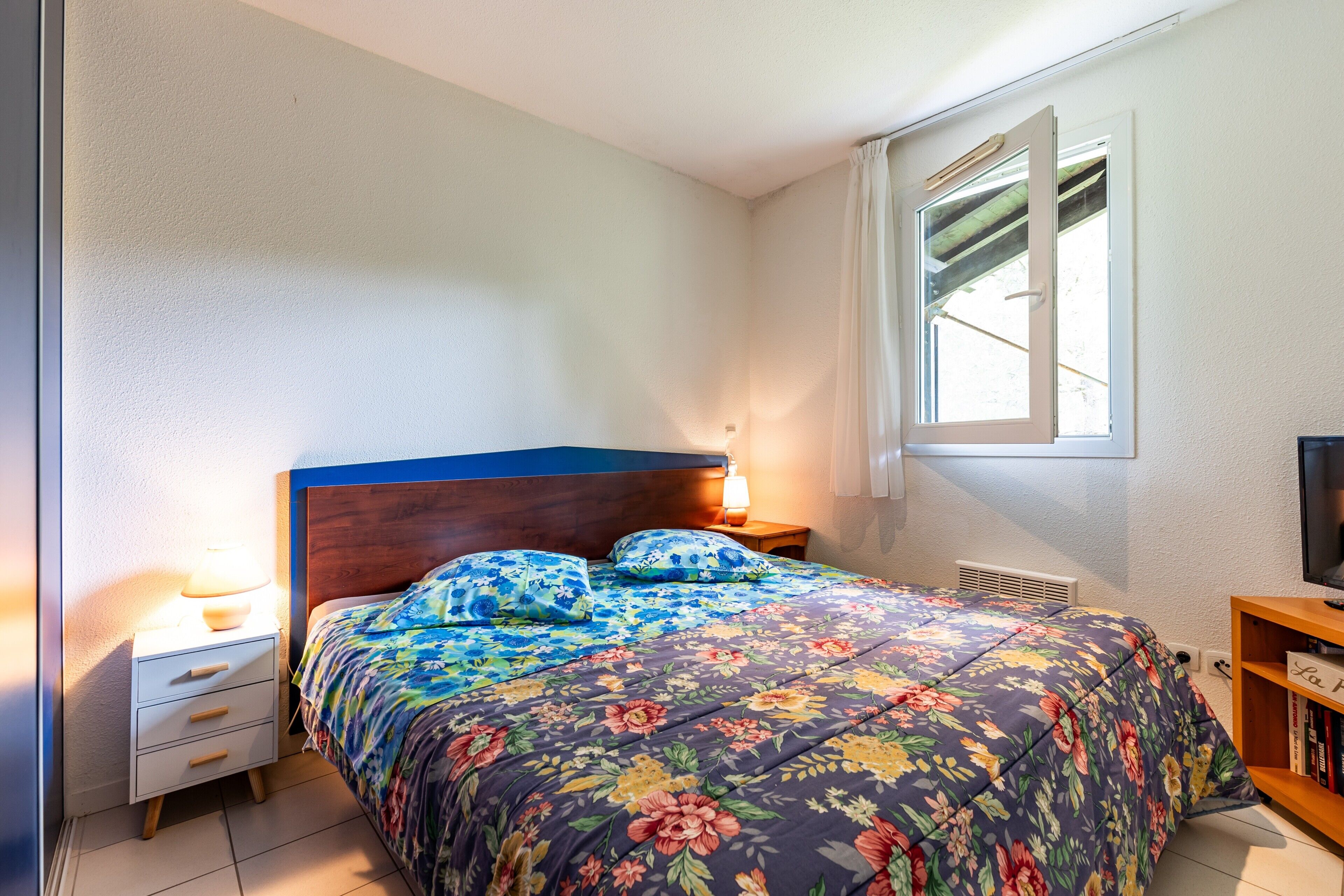 Een slaapkamer met een bed, nachtkastje, lamp, raam en een boekenkast.