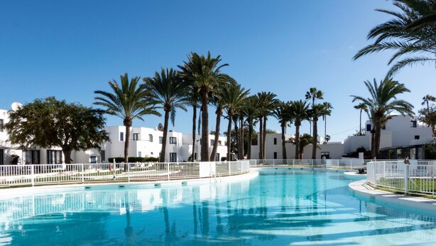 A swimming pool with a white fence, surrounded by palm trees and white buildings. - Holiday Home 'Los Barqueros 13' with Shared Pool, Private Terrace & Wi-Fi (Corralejo)