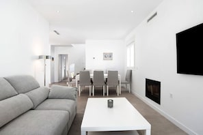 A modern living room with a sofa, a white coffee table, a dining area with chairs, and a fireplace.