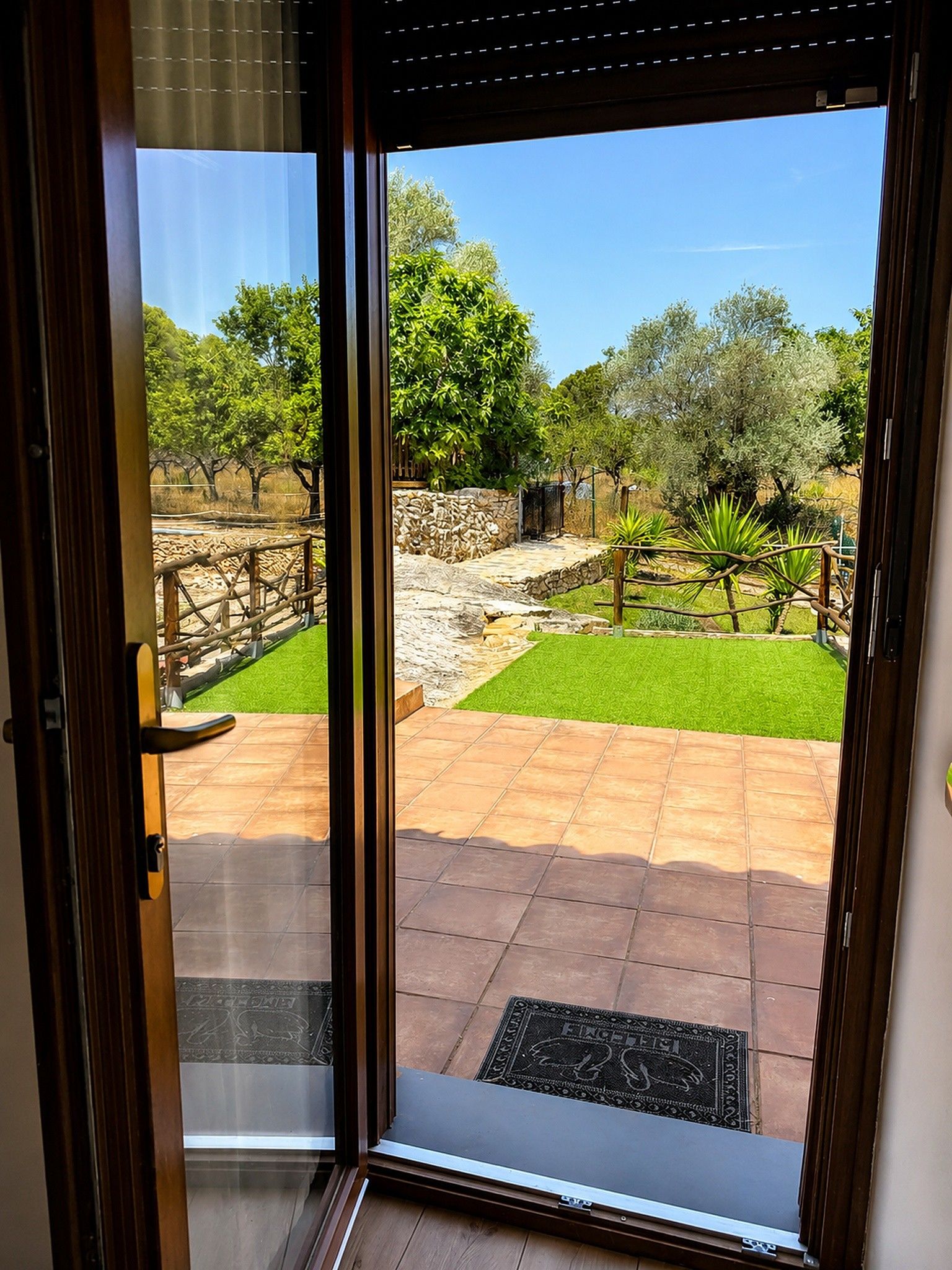 Vista a través de un ventanal a un patio con pared de piedra, valla de madera y césped.