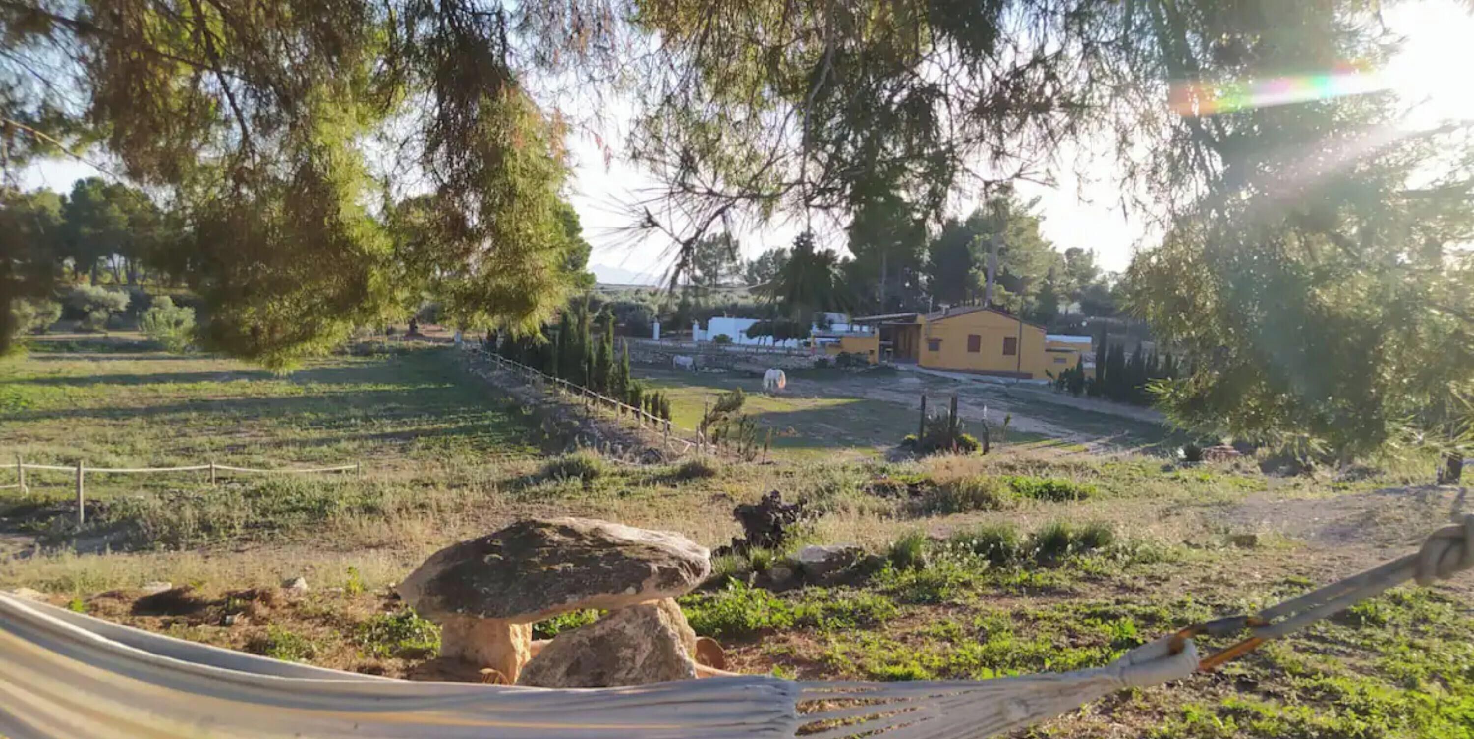 Paisaje rural con una hamaca, una construcción de piedra y un edificio amarillo a lo lejos.