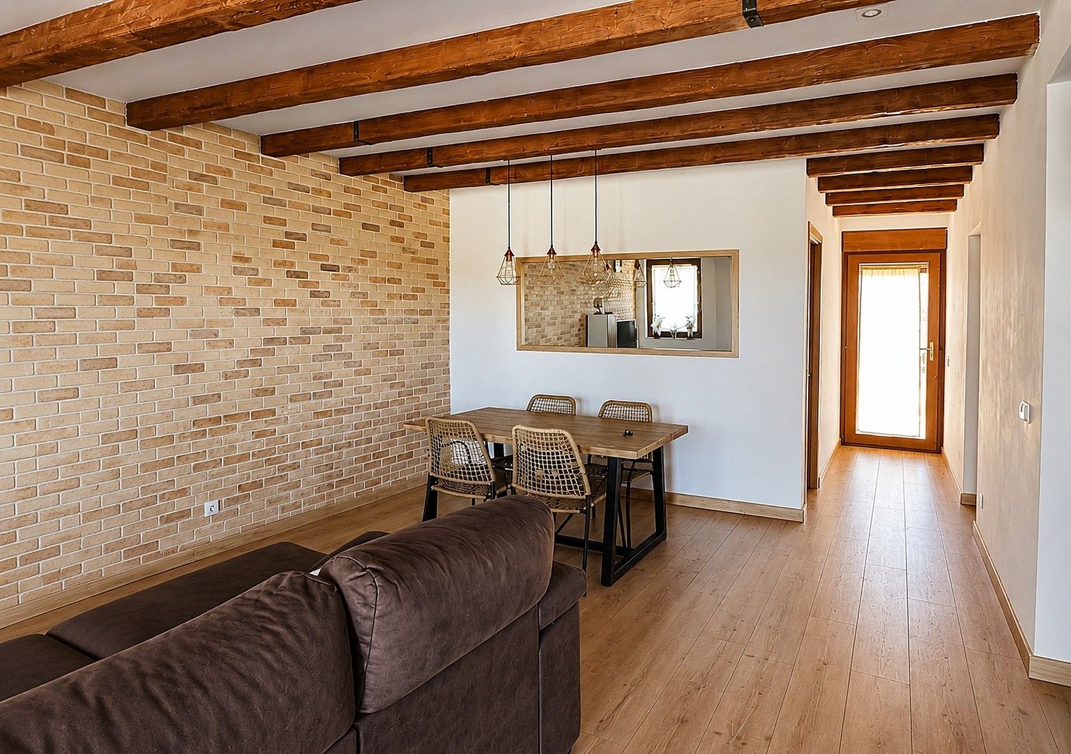 A dining area with a brick wall, wooden ceiling beams, a dining table with chairs, and a sofa.