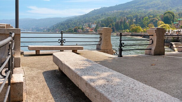 A stone bench and railing by a body of water with a view of a hillside town. - 'Casa Vacanza Malva' Apartment with Wi-Fi (Baveno)