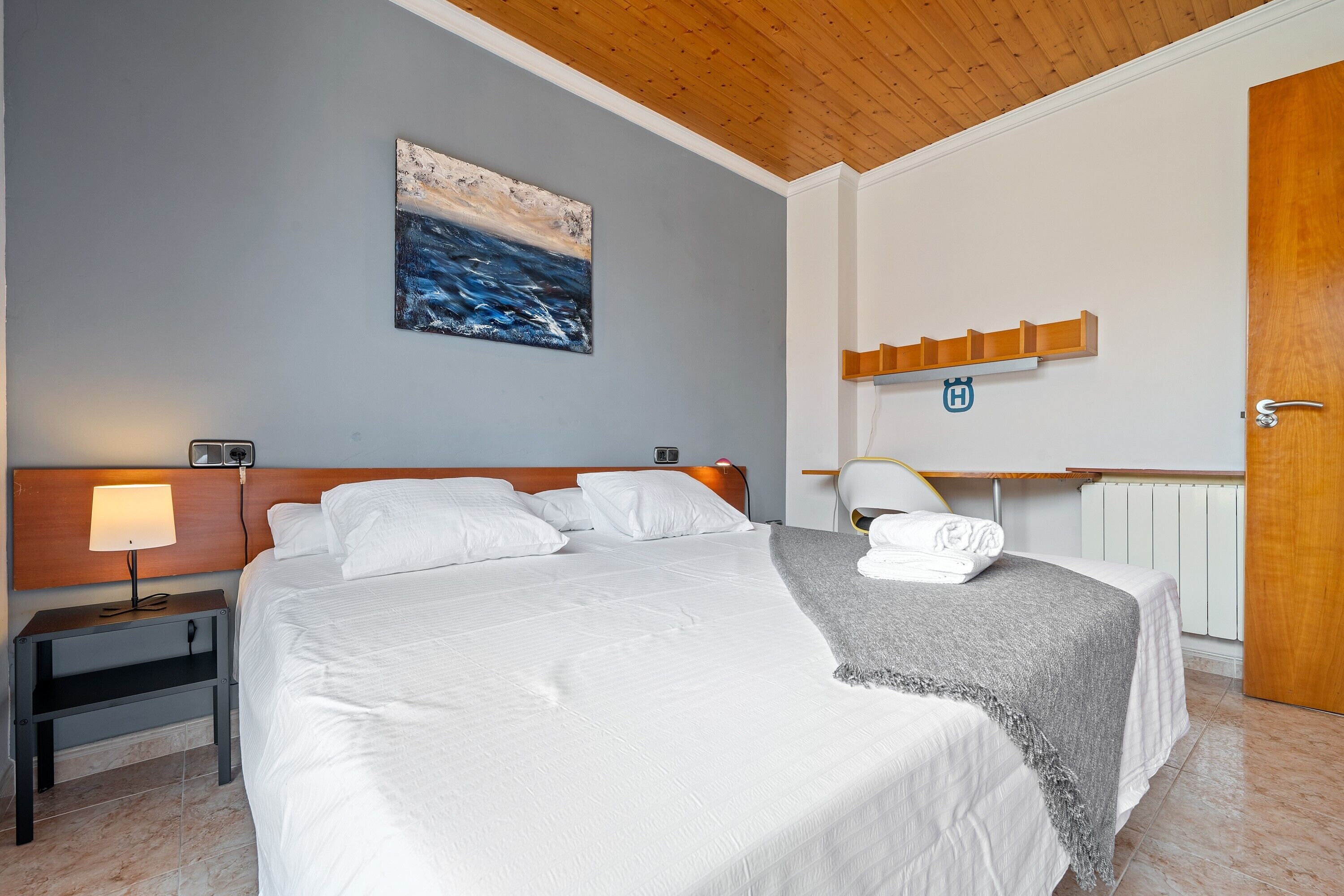 A hotel room with a bed, a nightstand, a lamp, a painting, a towel, a wooden shelf, and a door.
