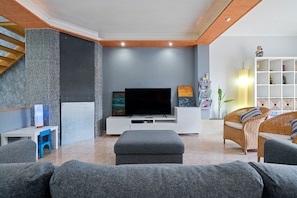 A modern living room with a flat-screen TV, grey sofas, a white entertainment unit, and a staircase with wooden railings.