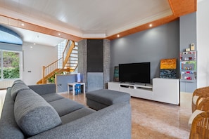 A modern living room with a grey sectional sofa, a flat-screen TV on a white stand, a staircase, and a bookshelf with various items.