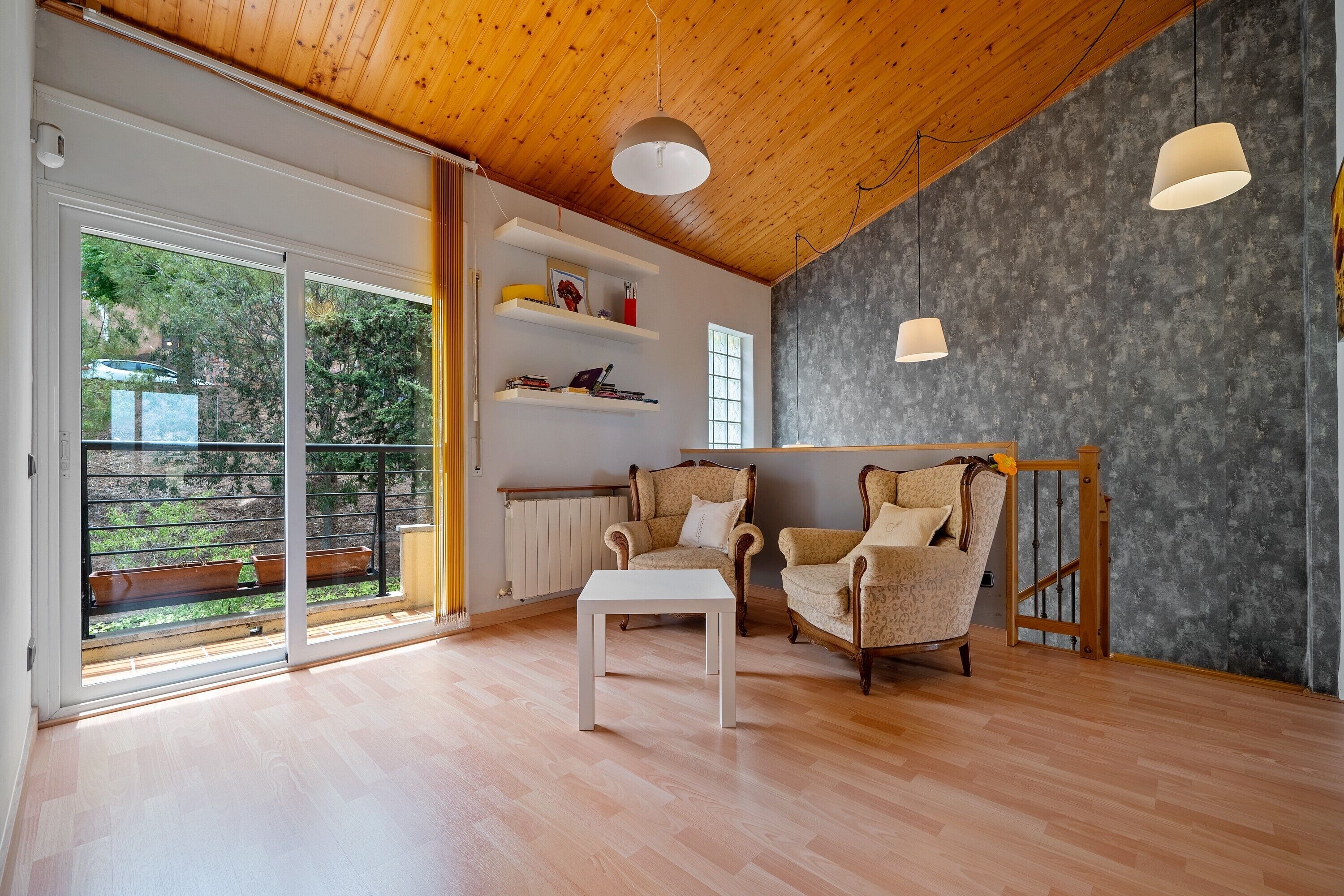 A spacious room with a wooden ceiling, a white table, two patterned armchairs, a bookshelf, and a view through a sliding glass door.