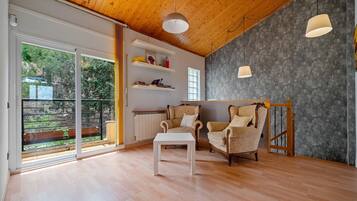 A spacious room with a wooden ceiling, a white table, two patterned armchairs, a bookshelf, and a view through a sliding glass door.
