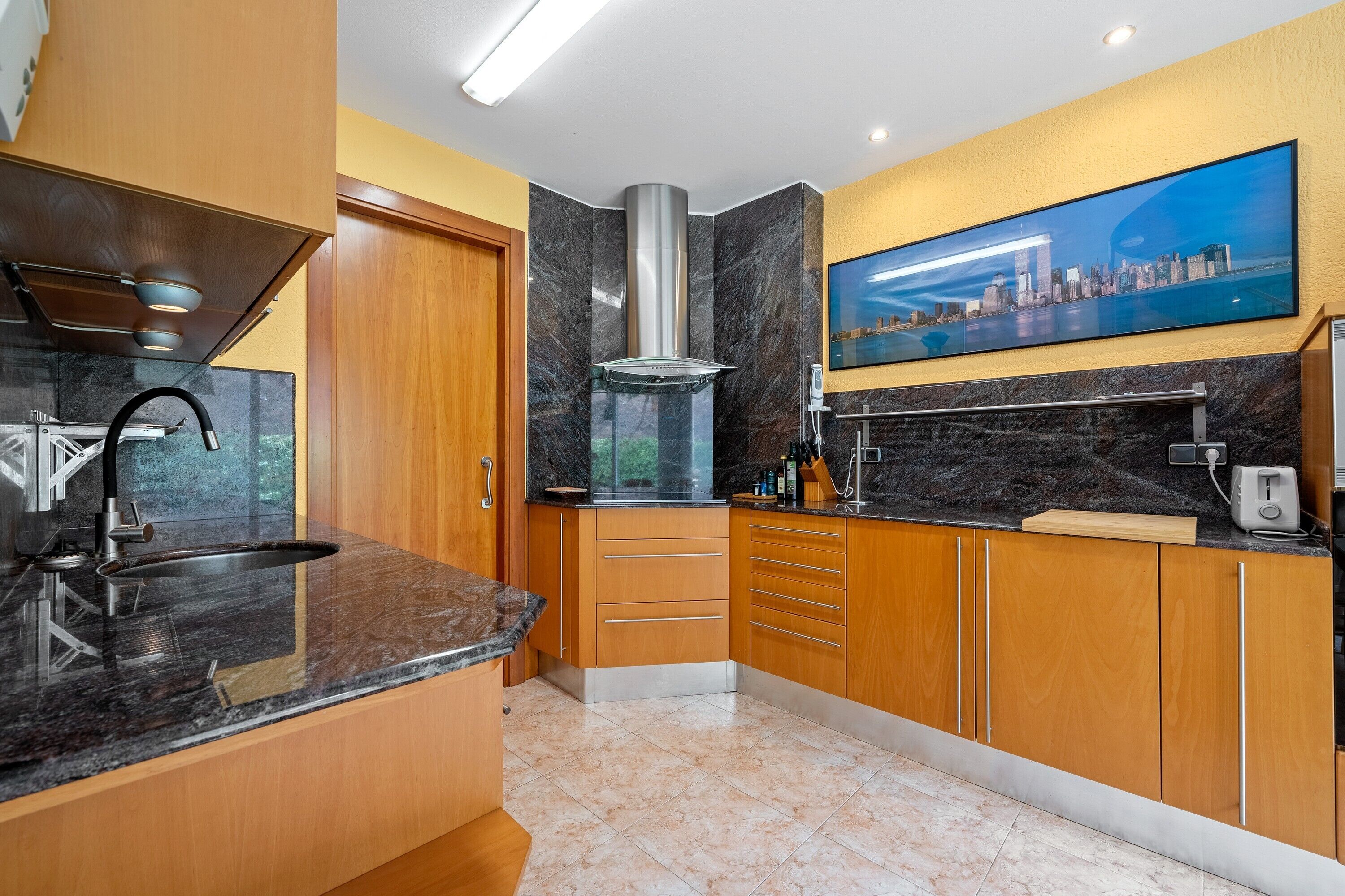 A modern kitchen with a built-in stove, a stainless steel range hood, wooden cabinets, a marble countertop, and a cityscape painting on the wall.