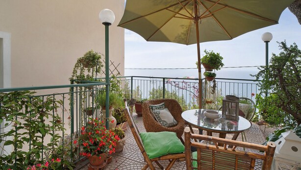 Een balkon met een tafel, stoelen, een parasol en bloempotten.