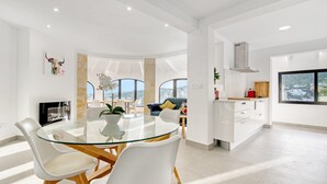 A modern living space with a glass dining table, white chairs, a sofa, a kitchen area, and large windows offering a city view. - Villa 'Ericito' with Private Pool, Wi-Fi and Air Conditioning (Dénia)