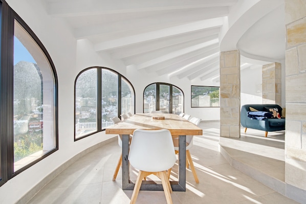 A modern dining area with a long table, white chairs, and large windows offering city views.