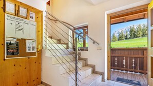 Un interno in legno con una scala, una bacheca con avvisi e vista su un paesaggio montano attraverso una porta.
