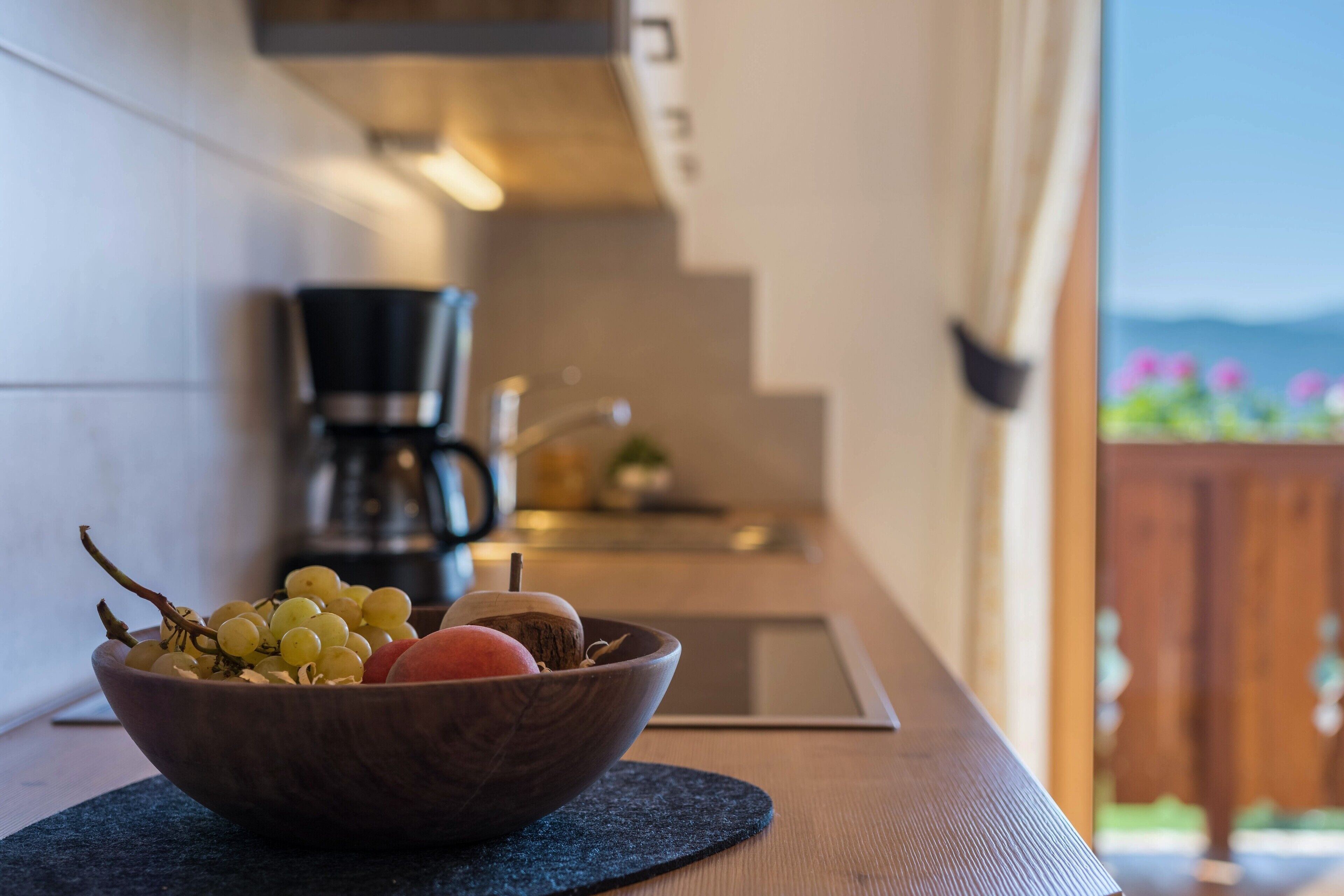 Un saladier en bois contenant des fruits, posé sur une assiette sombre, une machine à café, et une vue sur une terrasse en bois agrémentée de fleurs.
