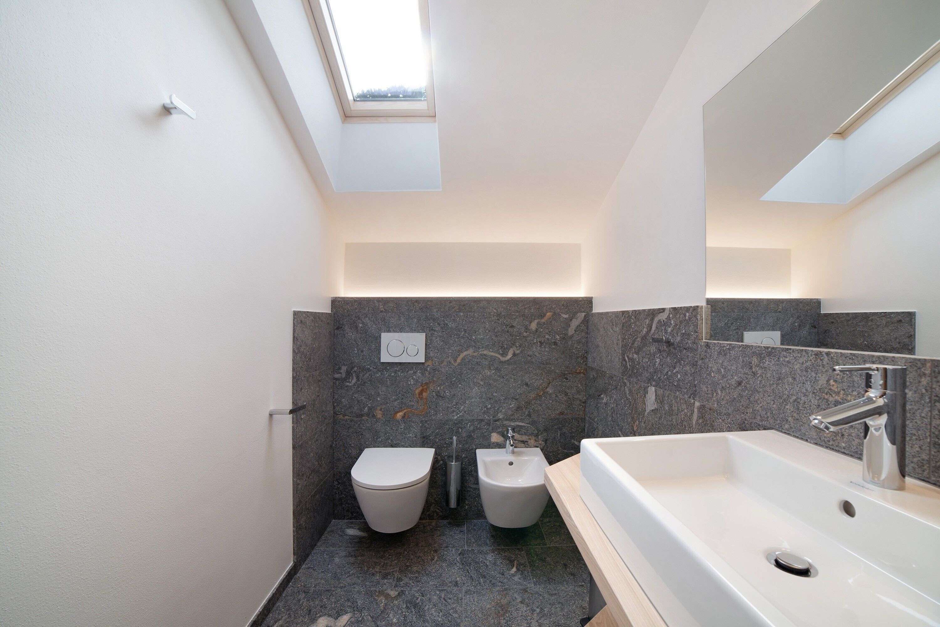 Een moderne badkamer met dakraam, een bidet, een wastafel en marmeren wanden.