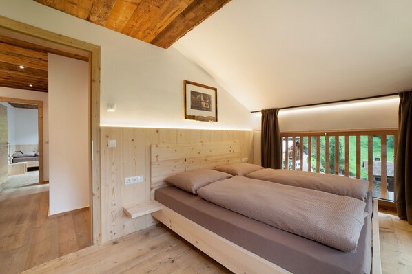A bedroom with a wooden headboard, a bed with beige bedding, a bedside table, and a window with a view of greenery.