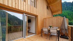 A wooden deck with a table and chairs, a sliding glass door, and a view of a forested hillside. - Apartment 'Herrischner 1' with Mountain View, Private Terrace and Wi-Fi (Moos in Passeier)