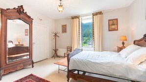 A bedroom with a bed, a wooden dresser, a mirror, a window with curtains, and a view of greenery. - La serre, grande maison  dans un grand jardin. (Laborde)
