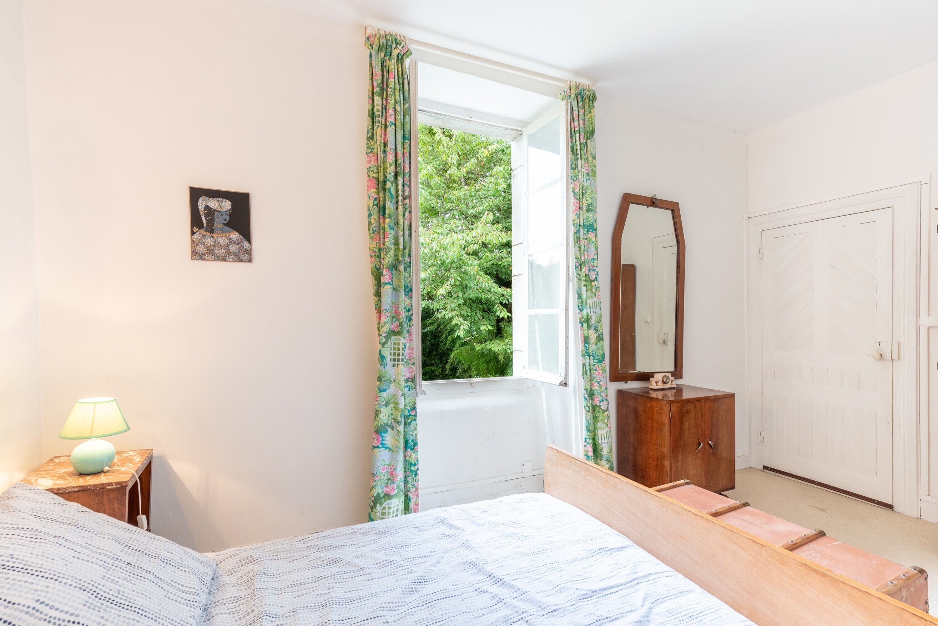 Een slaapkamer met een bed, een nachtkastje met lamp, een dressoir, een spiegel en een raam met gordijnen.