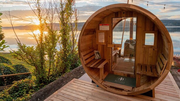 Une cabane en bois avec un banc et une table, située sur une terrasse en bois au bord d’un lac, avec un coucher de soleil en arrière-plan.