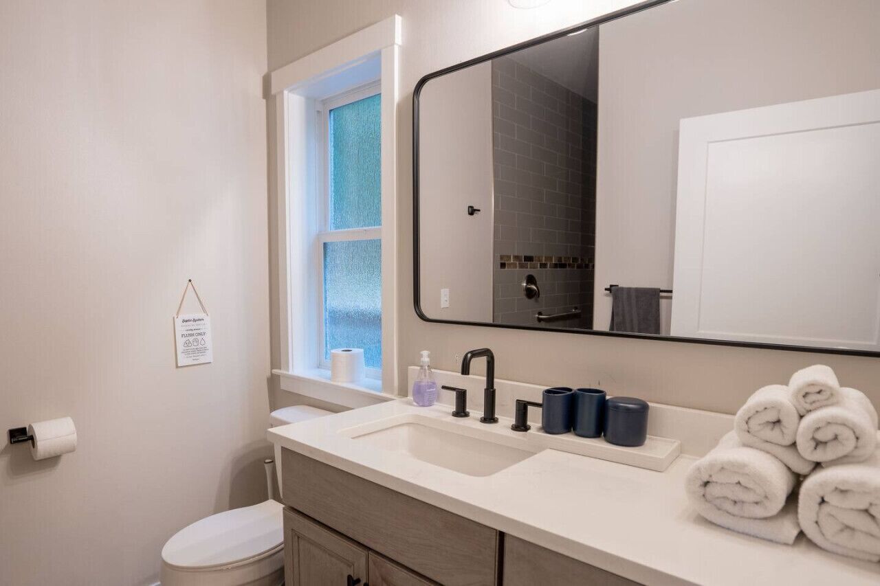 A modern bathroom with a white sink, black faucet, and a large mirror above it. There are towels stacked on the counter and a window with a view of the outside.