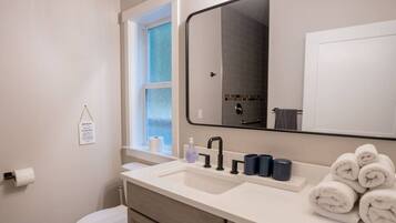 A modern bathroom with a white sink, black faucet, and a large mirror above it. There are towels stacked on the counter and a window with a view of the outside.