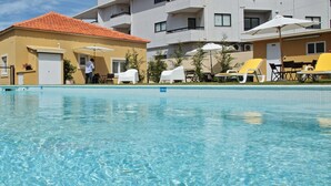Ein Schwimmbecken mit klarem Wasser, umgeben von weißen Liegestühlen und Sonnenschirmen, mit einem Gebäude mit Balkonen im Hintergrund.