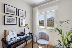Ein Zimmer mit Schreibtisch, gerahmten Bildern, einem Sessel, einem Fenster mit Blick auf Gebäude und einer Pflanze.