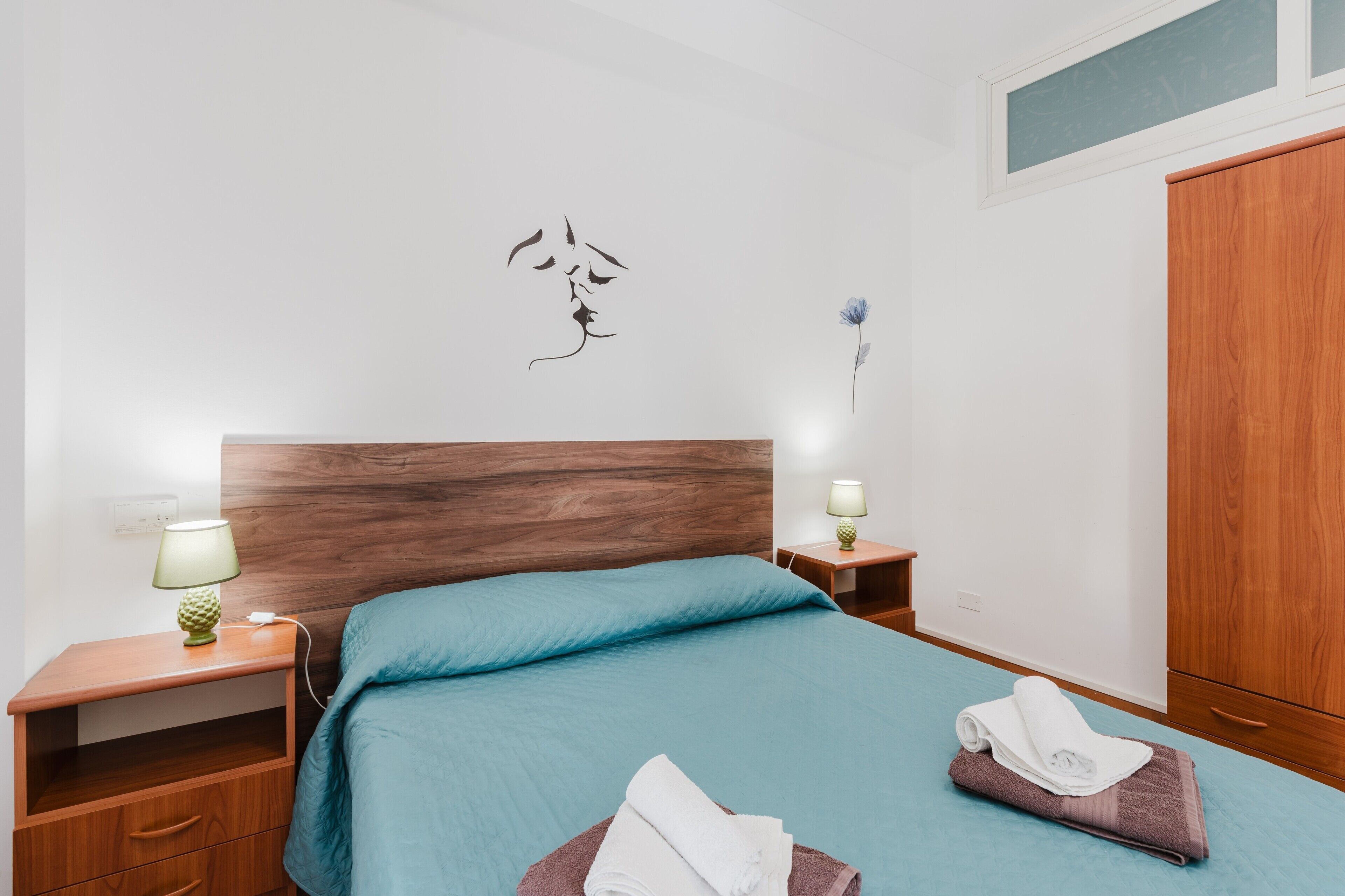 Un lit bien fait, recouvert d’une couvre-lit bleue, deux tables de chevet avec des lampes, une tête de lit en bois et une armoire.