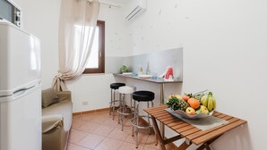 Eine kleine Küche mit Kühlschrank, einem Esstisch mit Hocker, einem Fenster mit Vorhängen und einer Wand-Klimaanlage.