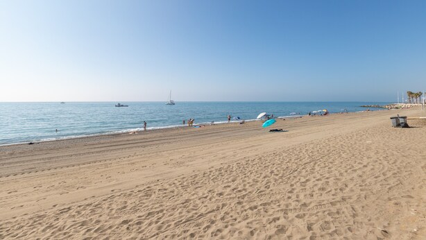 Une vaste plage de sable fin, un ciel bleu dégagé, une mer calme et quelques bateaux visibles au loin.