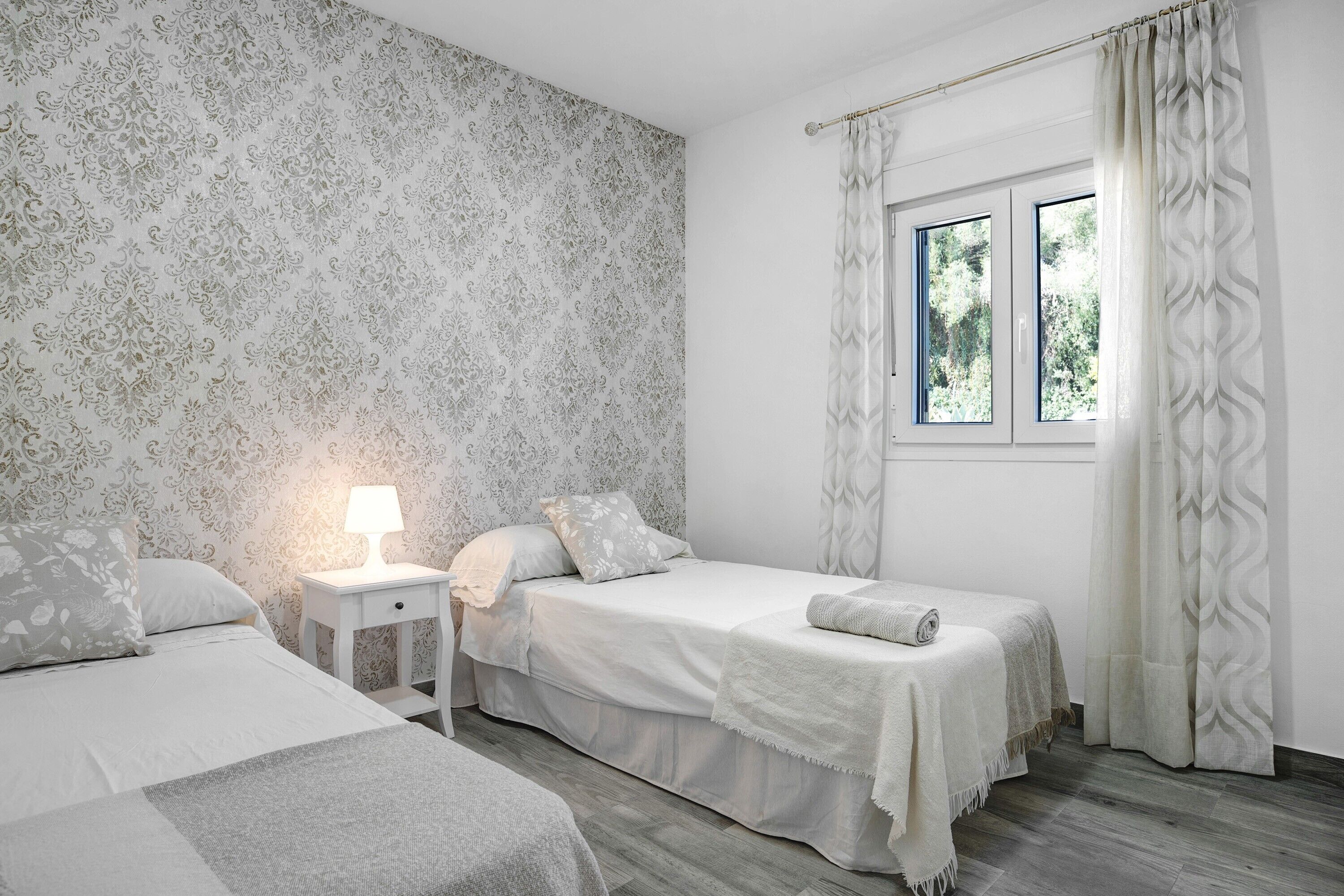 A bedroom with two beds, a nightstand, a window with curtains, and a patterned wallpaper.