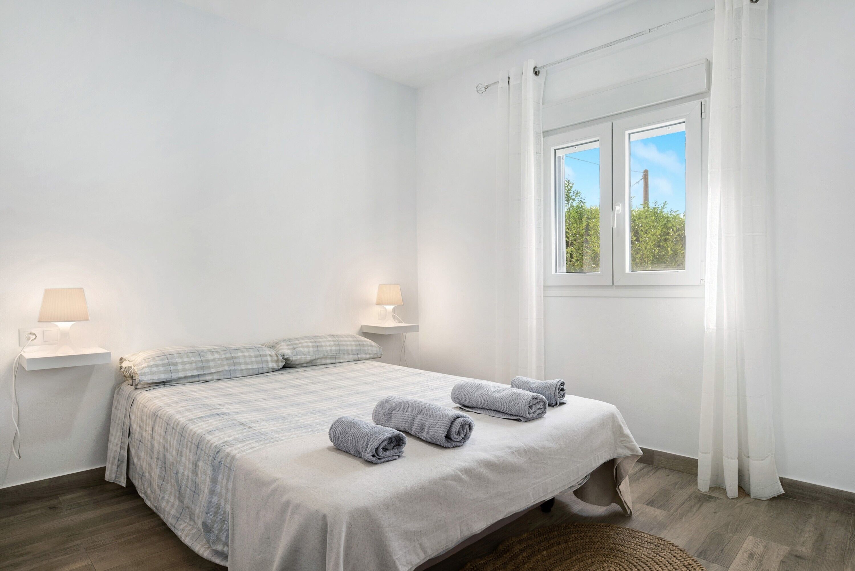 A bedroom with a bed, two bedside lamps, a window with curtains, and a view of greenery outside.