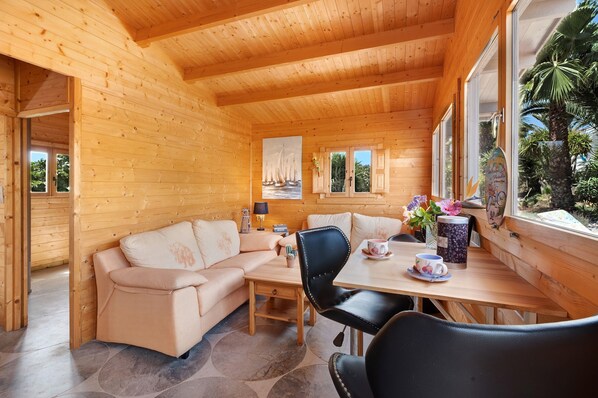 A wooden cabin interior with a sofa, dining table, chairs, and a window with a view of palm trees.