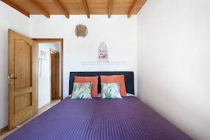 Une chambre avec une porte en bois, un lit recouvert d’une couverture violette, deux oreillers orange et une décoration murale représentant une planche de surf.