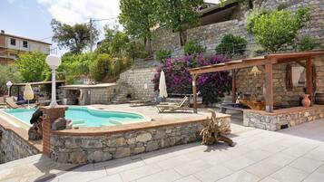 Un área de piscina con muros de piedra, sillas de descanso y una pérgola.
