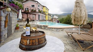 Un décor extérieur rustique avec une table en pierre, un tonneau en bois et une bouteille de vin. Au fond, on aperçoit une piscine et un bâtiment à la façade rose.