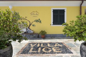 Une villa avec un mur jaune, une fenêtre à volets noirs et un paillasson en pierre sur lequel est inscrit « Villa Lice ».