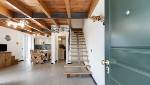 Un escalier en bois mène à une porte verte, on aperçoit un coin repas avec une table et des chaises en bois, et une cuisine au fond.