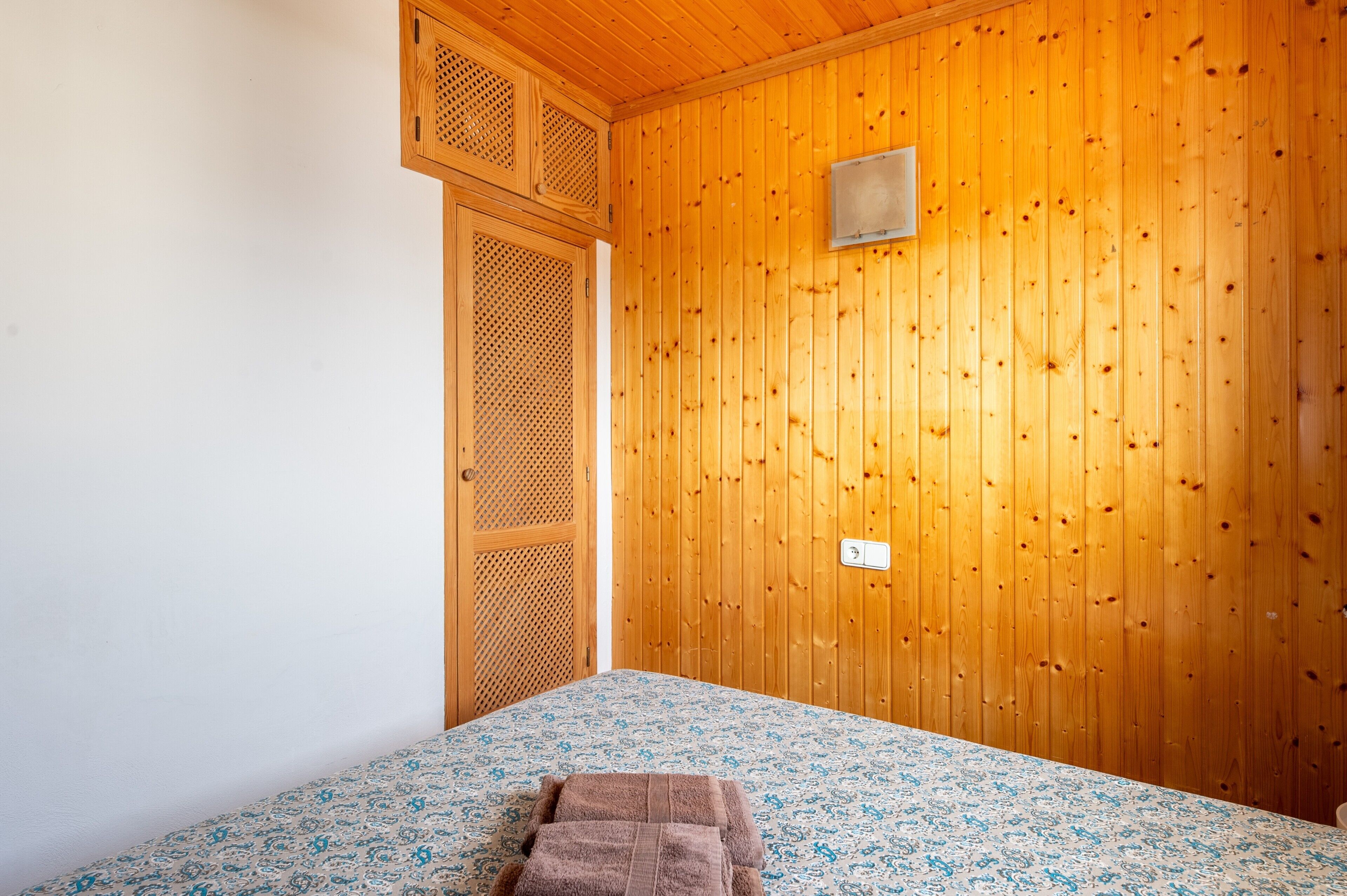 A room with wooden walls, a bed with a patterned bedspread, and a wooden door with a mesh screen.