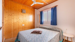 A room with wooden walls, a bed with a patterned bedspread, a ceiling fan, a window with curtains, and a small bedside table. - "Pujols 16" Apartment with a Balcony and Air Conditioning (Es Pujols)