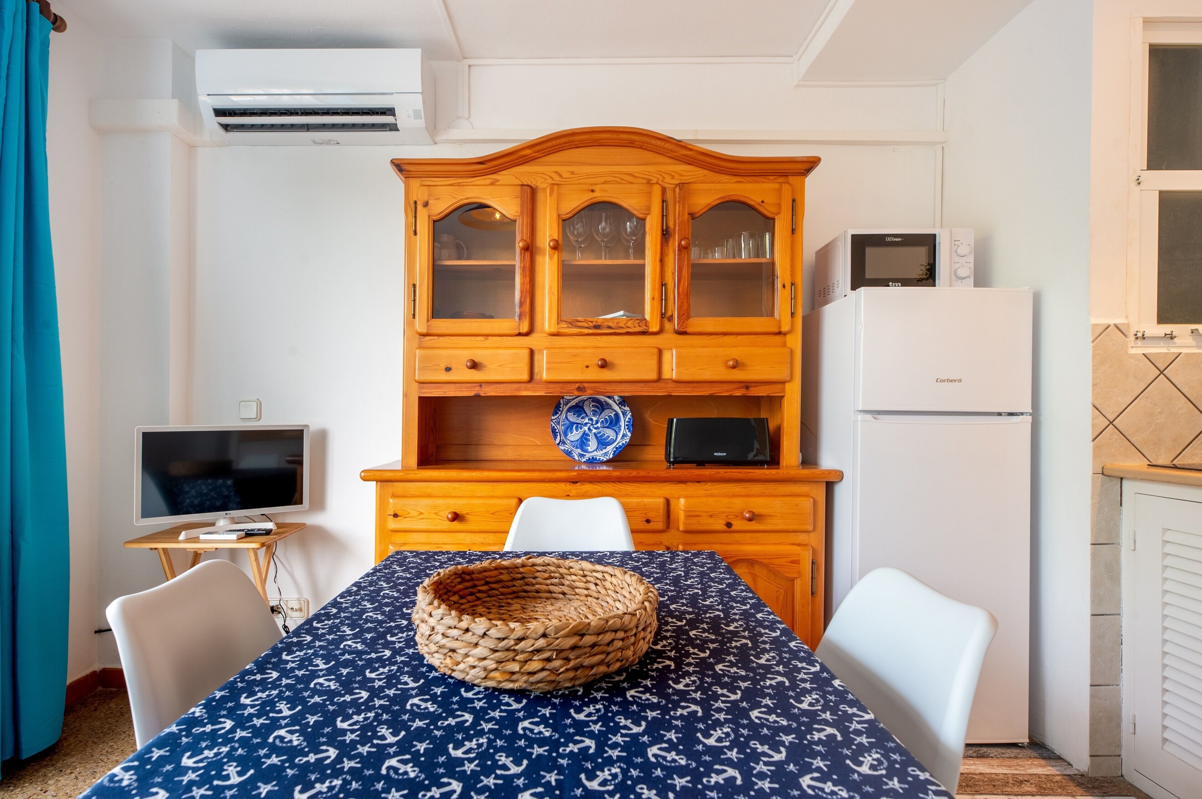 Une pièce de taille modeste comprenant une table à manger, des chaises, un téléviseur, un meuble avec des portes en verre, un four à micro-ondes et un réfrigérateur.