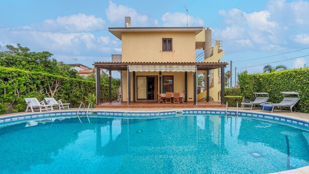 A swimming pool with lounge chairs, a covered patio, and a two-story building with a tiled roof.