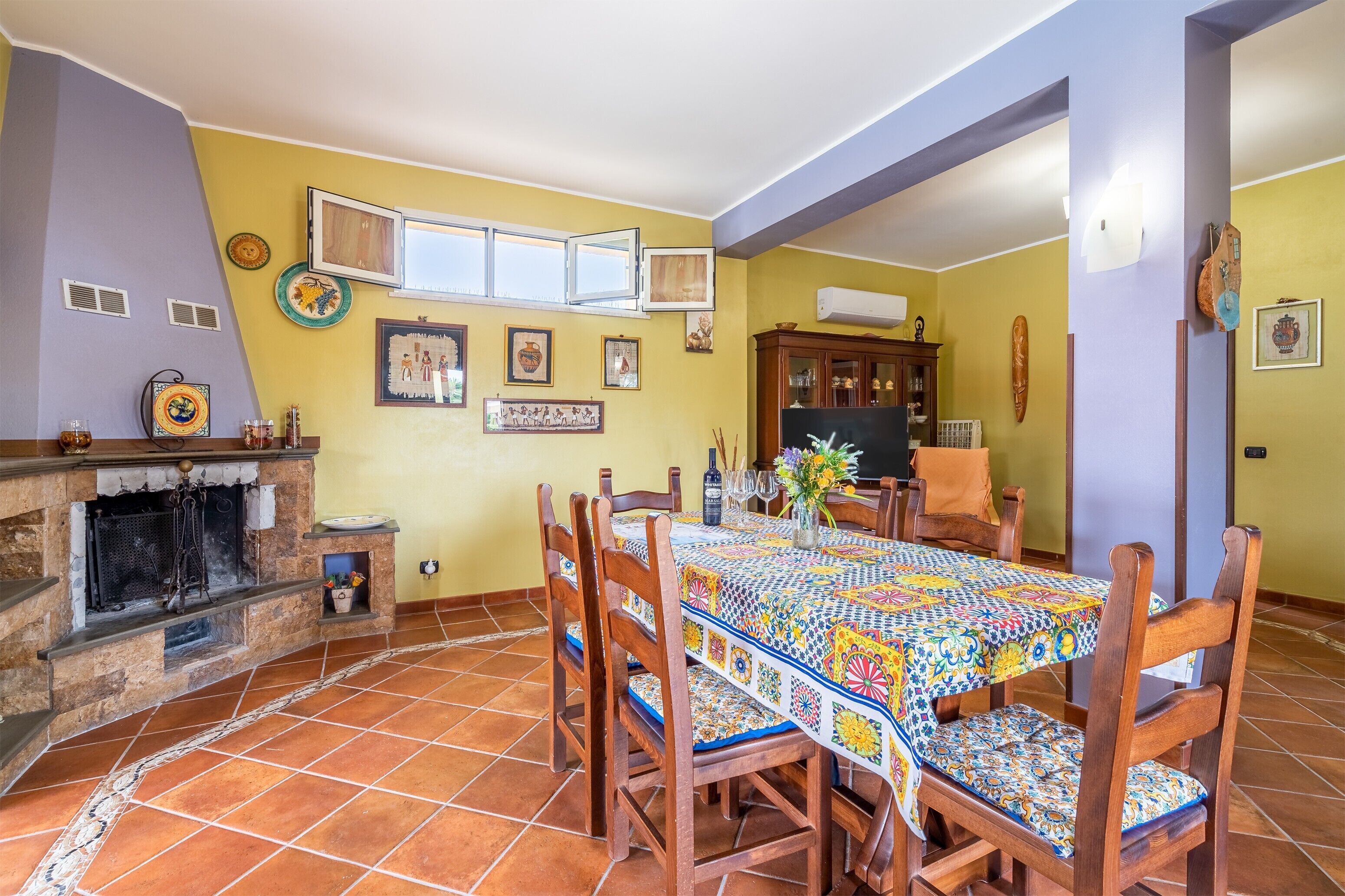 Uma área de refeições com piso ladrilhado, cadeiras de madeira, mesa com toalha colorida, lareira e decorações nas paredes.