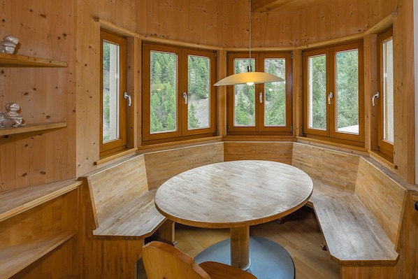 A wooden interior with a round table and benches, large windows with forest views, and a hanging light fixture.