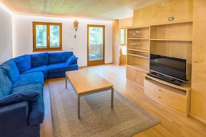 A living room with a blue sofa, a wooden coffee table, a TV on a wooden stand, and a view through a glass door.